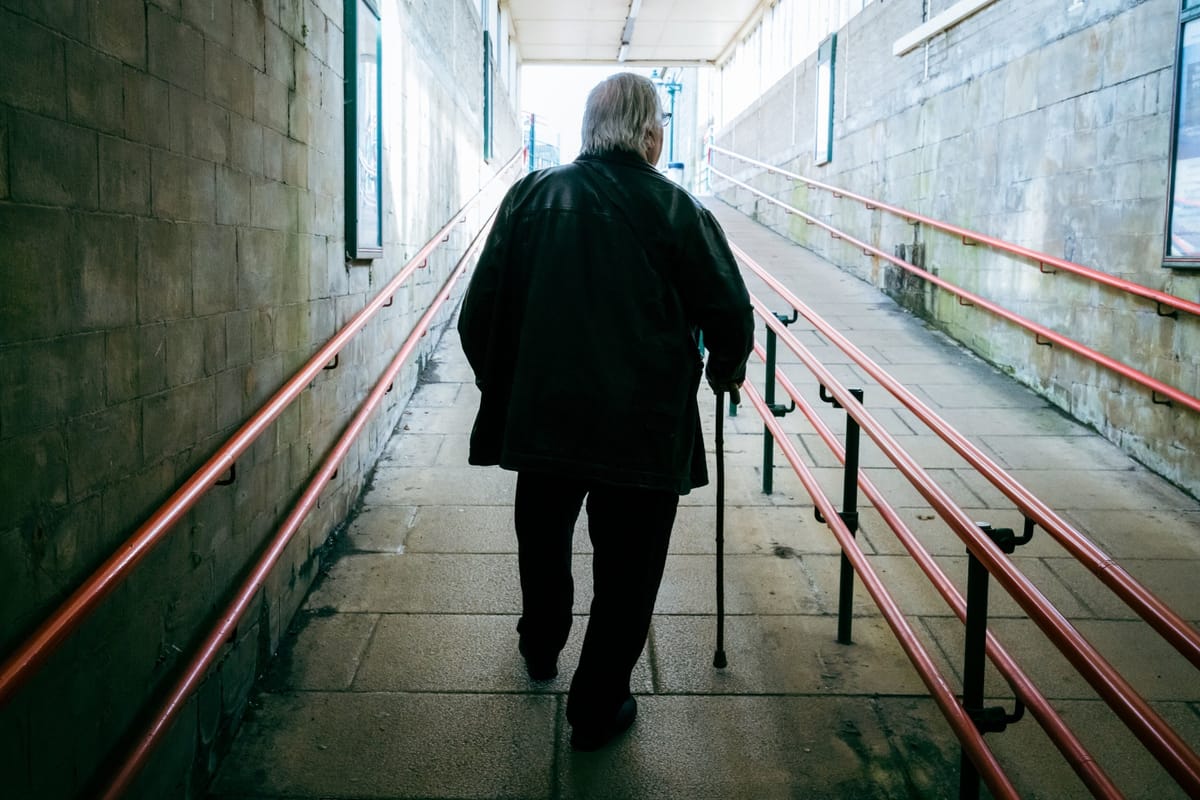 An old man with a cane in his right hand walks forward and upward to the outside, through a tunnel with a flat surface and many railings for accessibility.