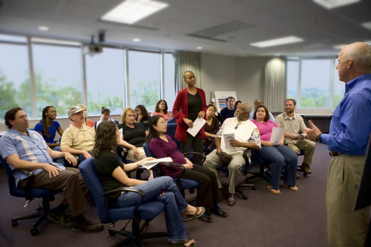 In an office space on an upper floor, in the day with trees outside the windows, a woman stands up during a meeting to address the presenter.