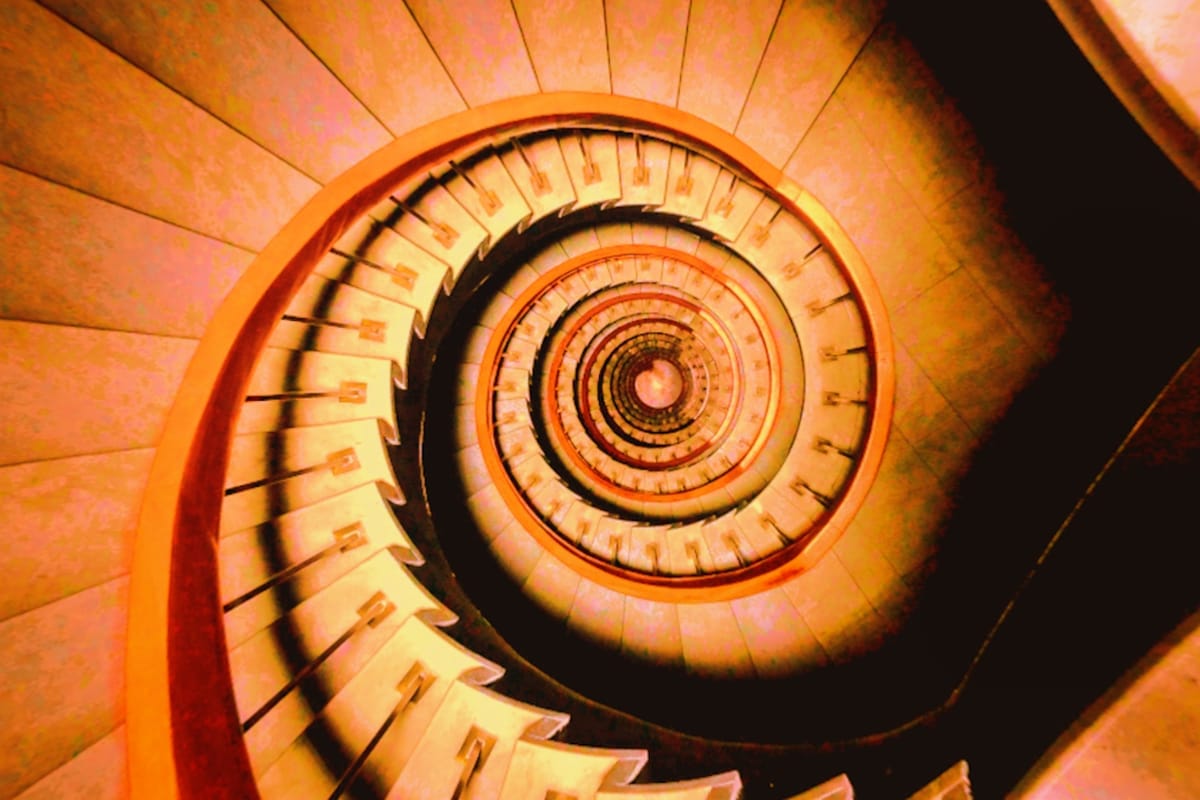 A red-tinged photograph looking down a spiral staircase of marble steps, brass and iron posts, and a smooth wooden railing