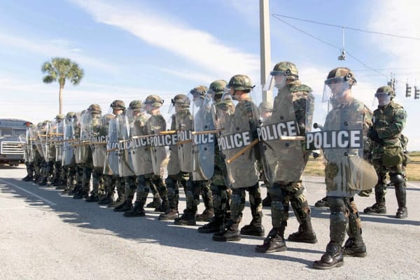 Soldiers in police riot gear train on a large road in Florida with a sunny sky and no shade, with one palm tree in the background. A traffic light is in the background.