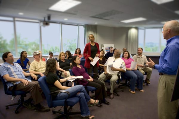In an office space on an upper floor, in the day with trees outside the windows, a woman stands up during a meeting to address the presenter.
