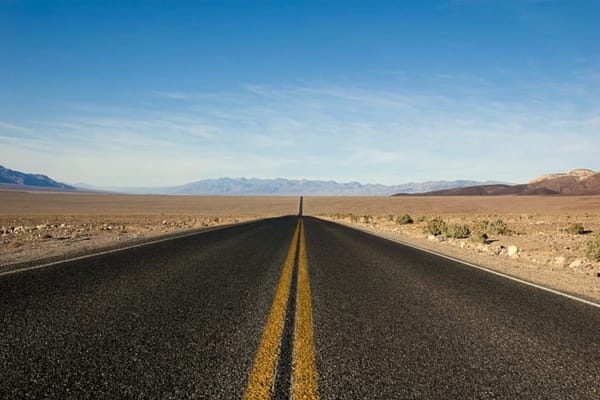 A two-lane road with a double yellow centerline that runs through a desert that is mostly flat, with an orange-brown-tinge on a sunny day with mountains in the far background
